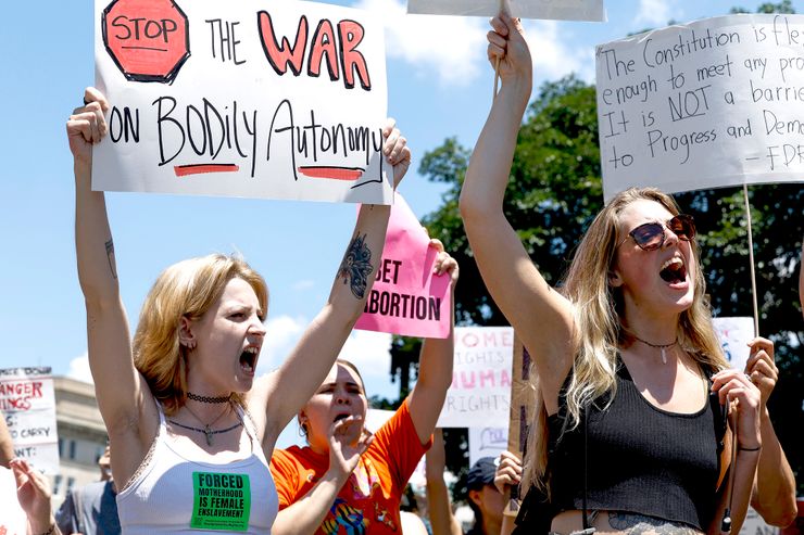 A White woman, wearing a white tank top and a label that reads "Forced motherhood is female enslavement," yells while holding a sign that reads, "Stop the war on bodily autonomy." People are protesting in the background. 