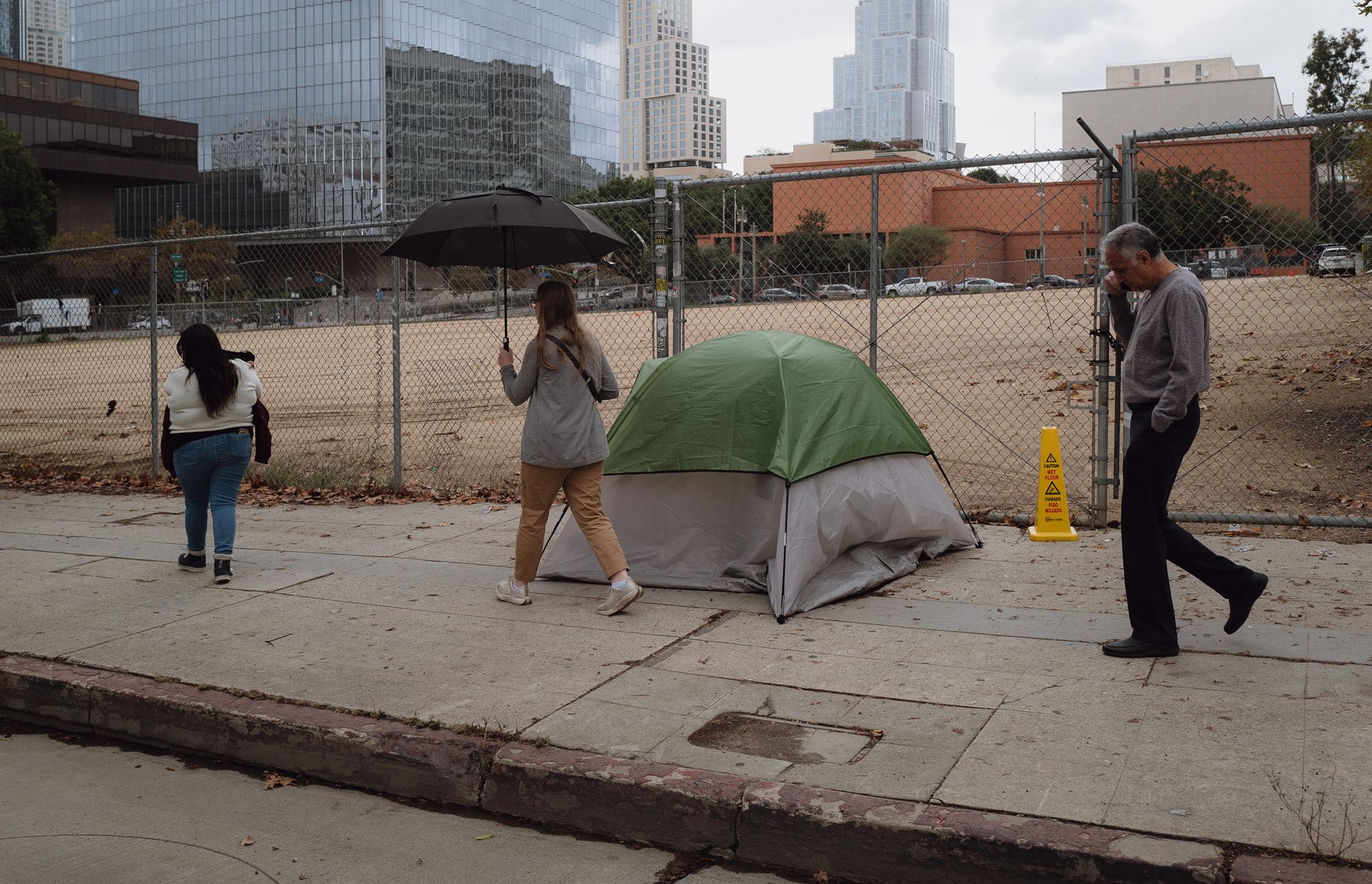 A photo shows Jazmine Mapes, a Native American woman with long dark hair, wearing a white sweater and jeans, walking near a chain link fence. An empty lot is situated behind the fence. In front of the fence is a tent. A woman holding an umbrella walks behind Mapes, and a man talking on a cell phone walks behind her.