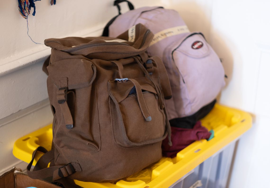 A brown backpack and a lilac backpack sit on a plastic tub with a yellow lid.