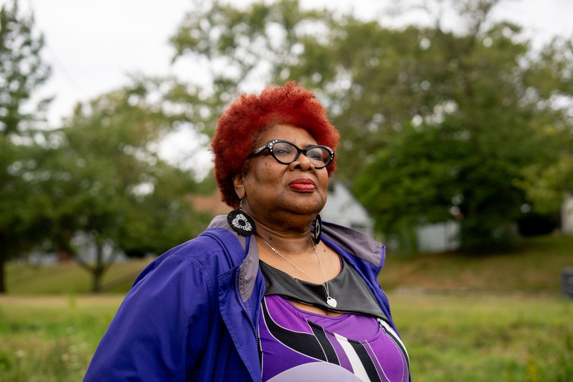 A Black woman with red hair stands outside in a grassy area with trees behind her.