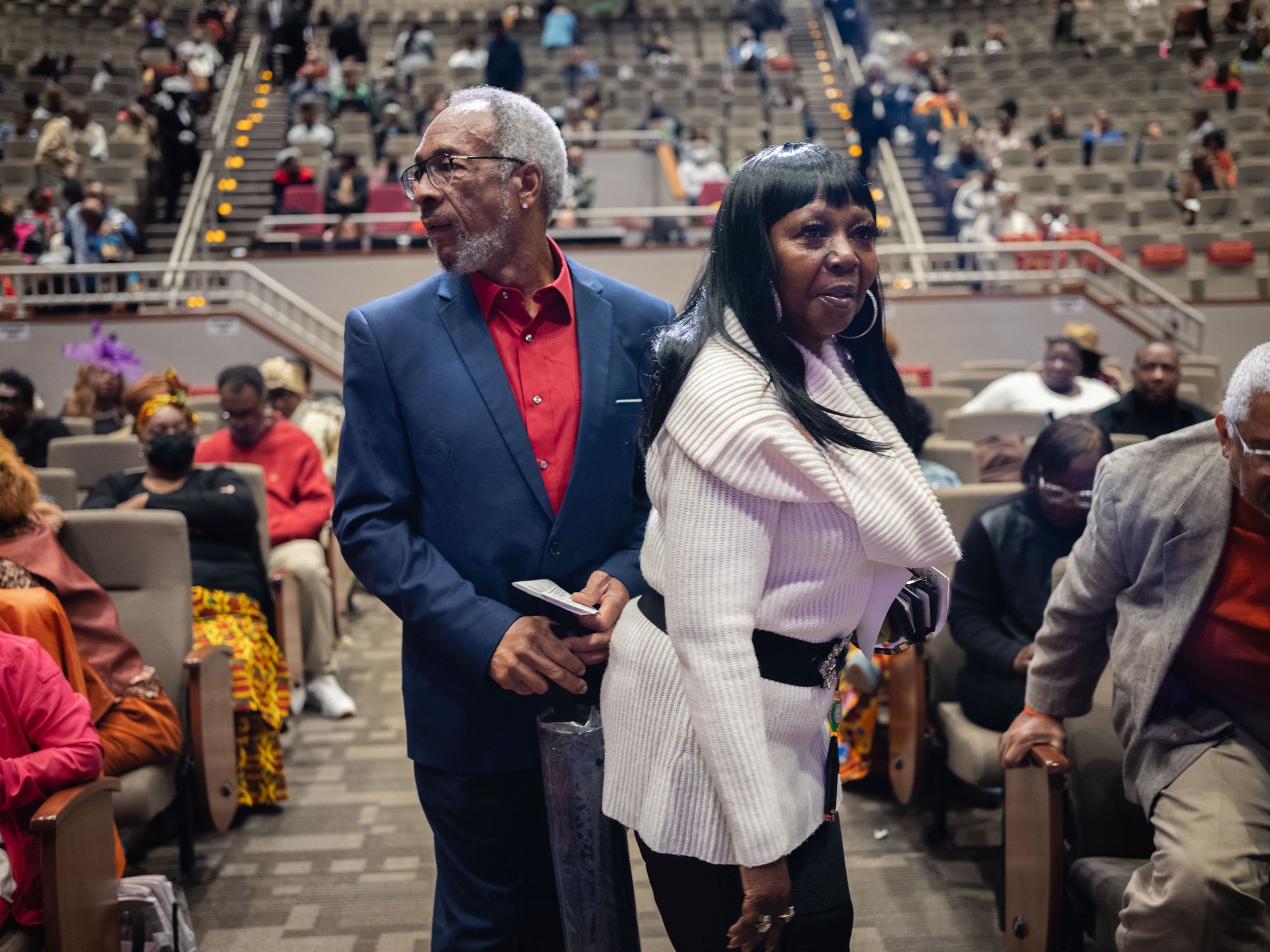 Moses, a Black man wearing a navy suit, and Armour, a Black woman wearing a white sweater, stand in the middle of an aisle in a church with rows of seats partially occupied by people.