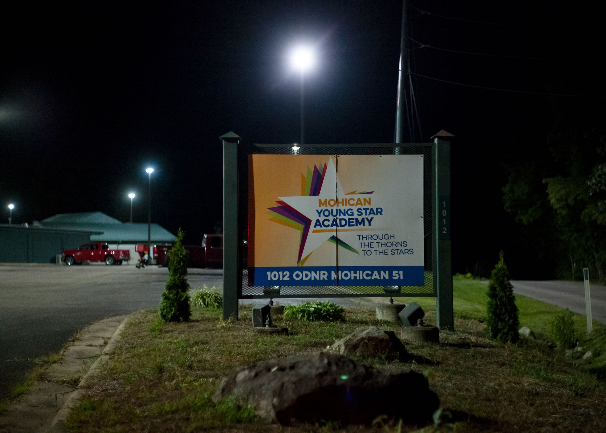 A photo shows a multicolored sign with a layered star logo, at night. The sign reads “Mohican Young Star Academy” and “Through the Thorns to the Stars.” Behind the sign is a parking lot with streetlights. 