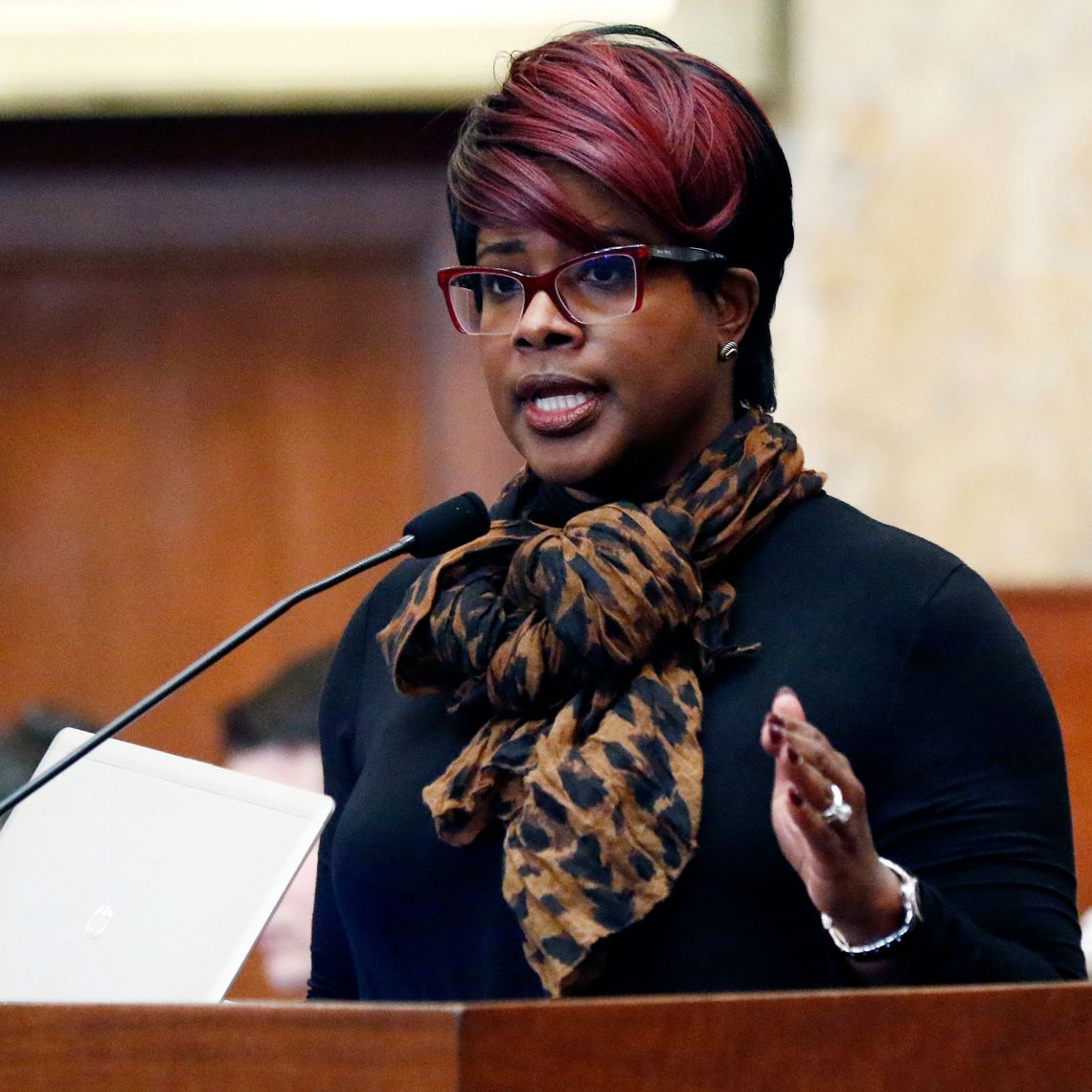 A Black woman with short hair with red bangs, wearing red framed glasses and a patterned scarf, speaks in front of a podium. 