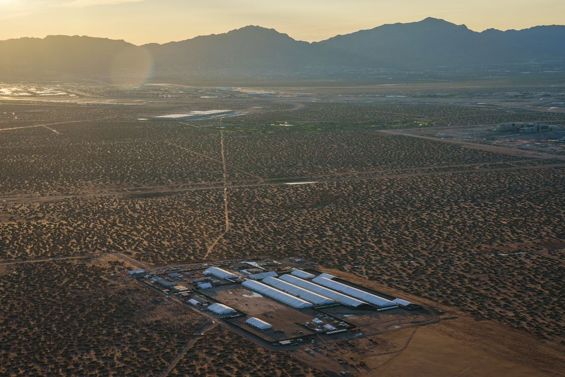 An aerial view shows a campus of long, white tents in a desert with a mountain range in the distance. 