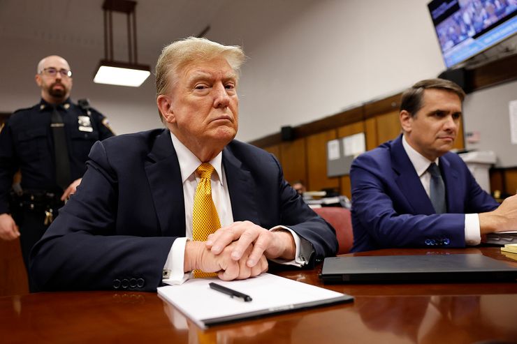 Former President Donald Trump, wearing a yellow tie with a dark-red graphical pattern, sits at a courtroom table, his hands folded, looking into the camera, while his defense attorney sits alongside him and a court officer stands watch behind them.  
