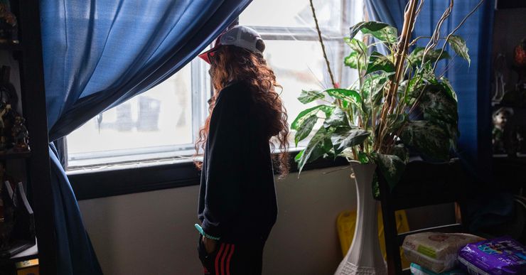 A woman with long, wavy red hair, whose face is not visible, looks out a window with blue curtains. Behind her is a tall vase with green leaves in it. A stack of diapers is on a chair.  