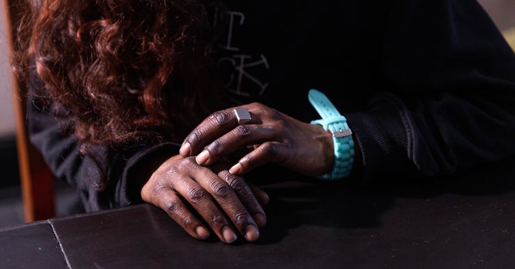 A photograph shows a closeup of a Black woman's hands. She wears a silver ring on the middle finger of her left hand, and an aqua-colored watch.  