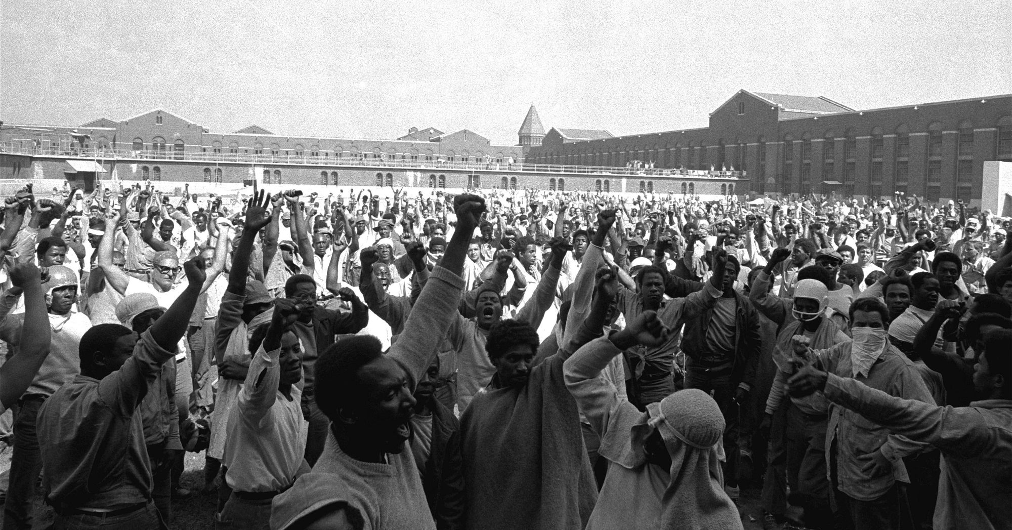A black-and-white photograph shows a large group of men in prison uniforms, a majority of whom are Black, with their fists up as they stand in a yard in front of a prison facility. 