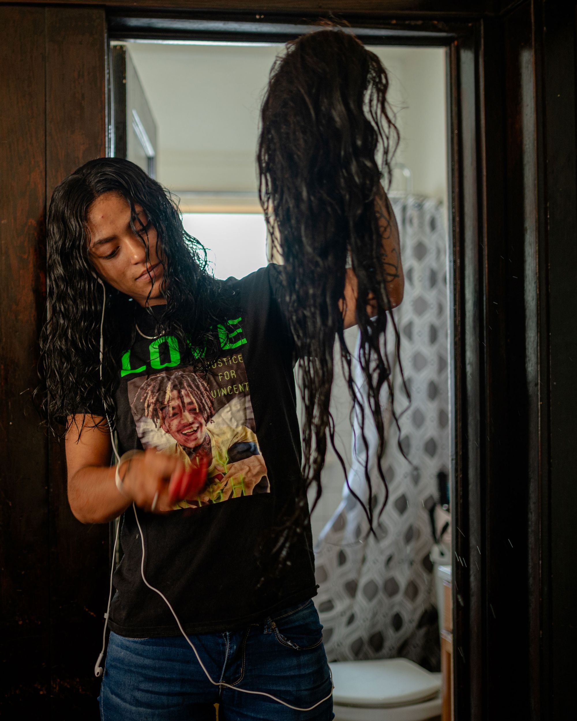 A photo of a young Black woman, wearing a shirt with a photo of a Black man. The woman is holding up and brushing a wig.
