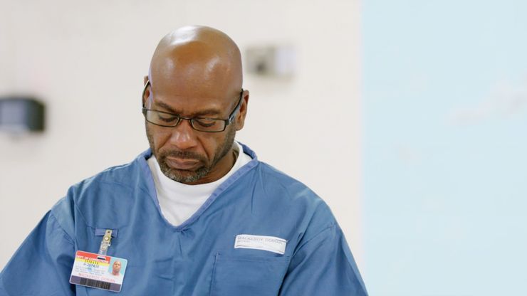 A Black man with square-framed glasses and a thin, slightly graying beard, sits in a Florida prison wearing a blue uniform and looking down.  