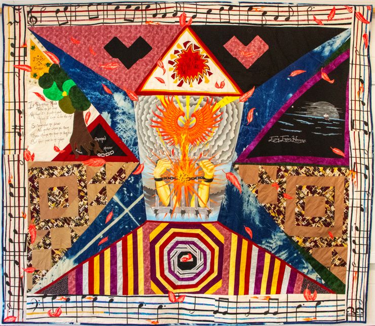 A photo of a quilt shows, at center, two arms with raised fists in handcuffs reaching out from behind prison bars. There is a phoenix rising from the center of the handcuffs, where there is a starburst. Multi-colored pieces surround the center, and the edges of the quilt are lined with a pattern of musical notes on a staff.
