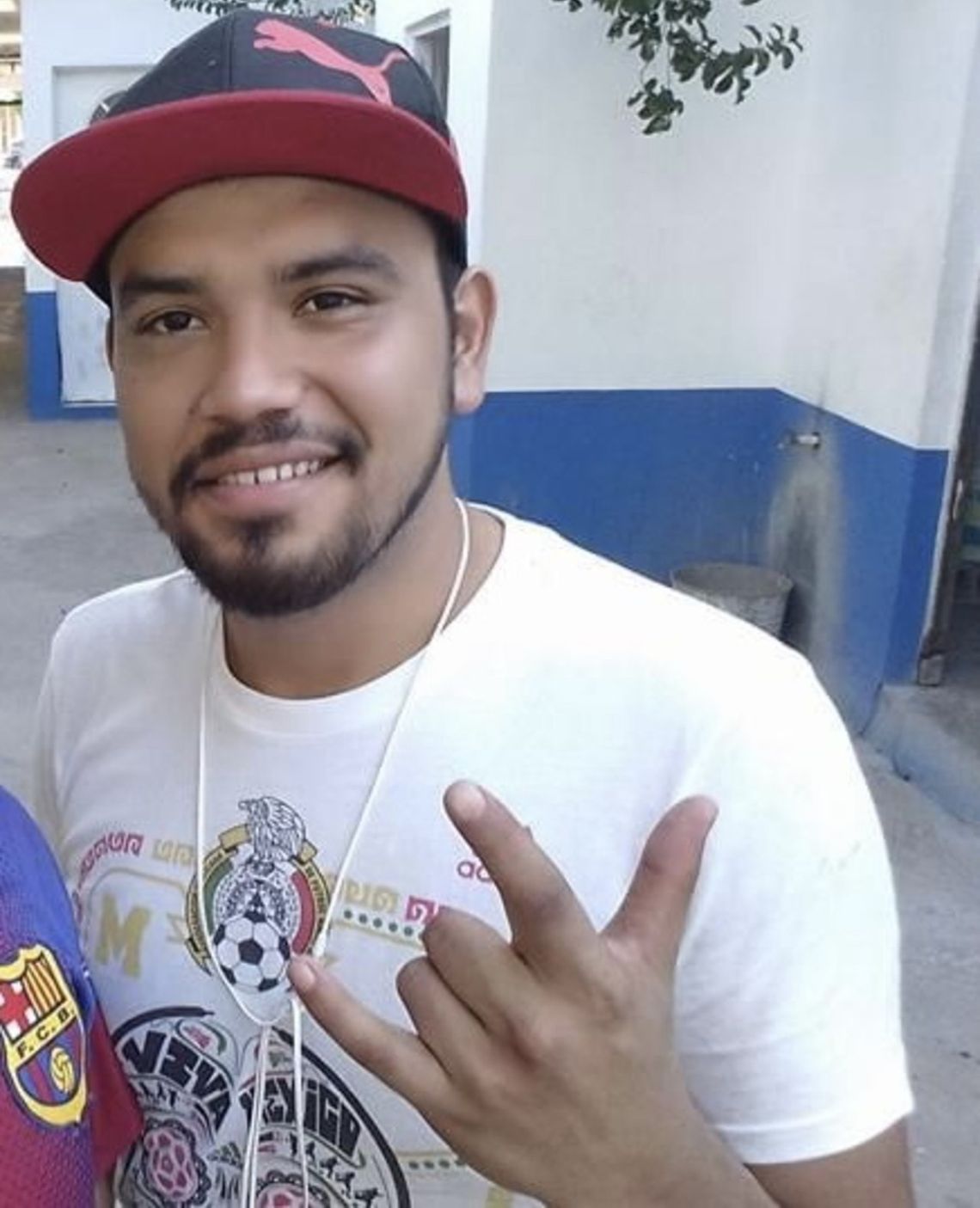 A man with medium-toned skin and a short beard wears a white t-shirt and a black baseball cap. 