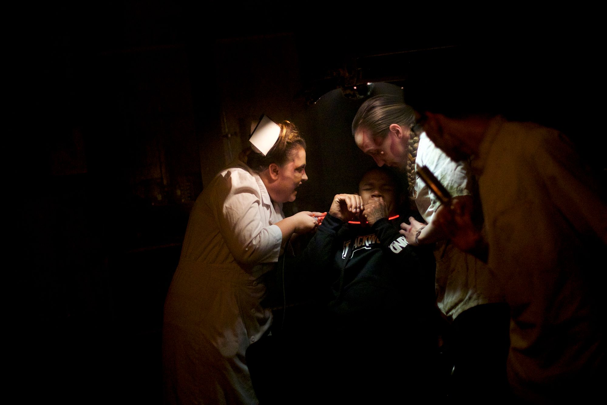 A woman dressed in a white nurse's uniform holds up a drill to a vistor's mouth, while he is being held down by another actor. 