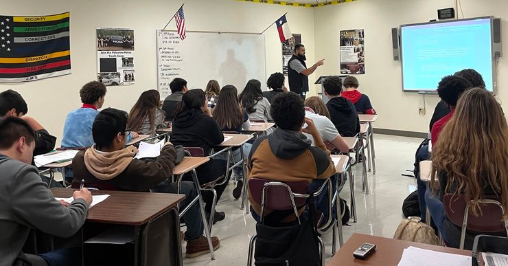 An instructor with a long black beard looks at a screen as he lectures in a classroom filled with students sitting in their desks. 