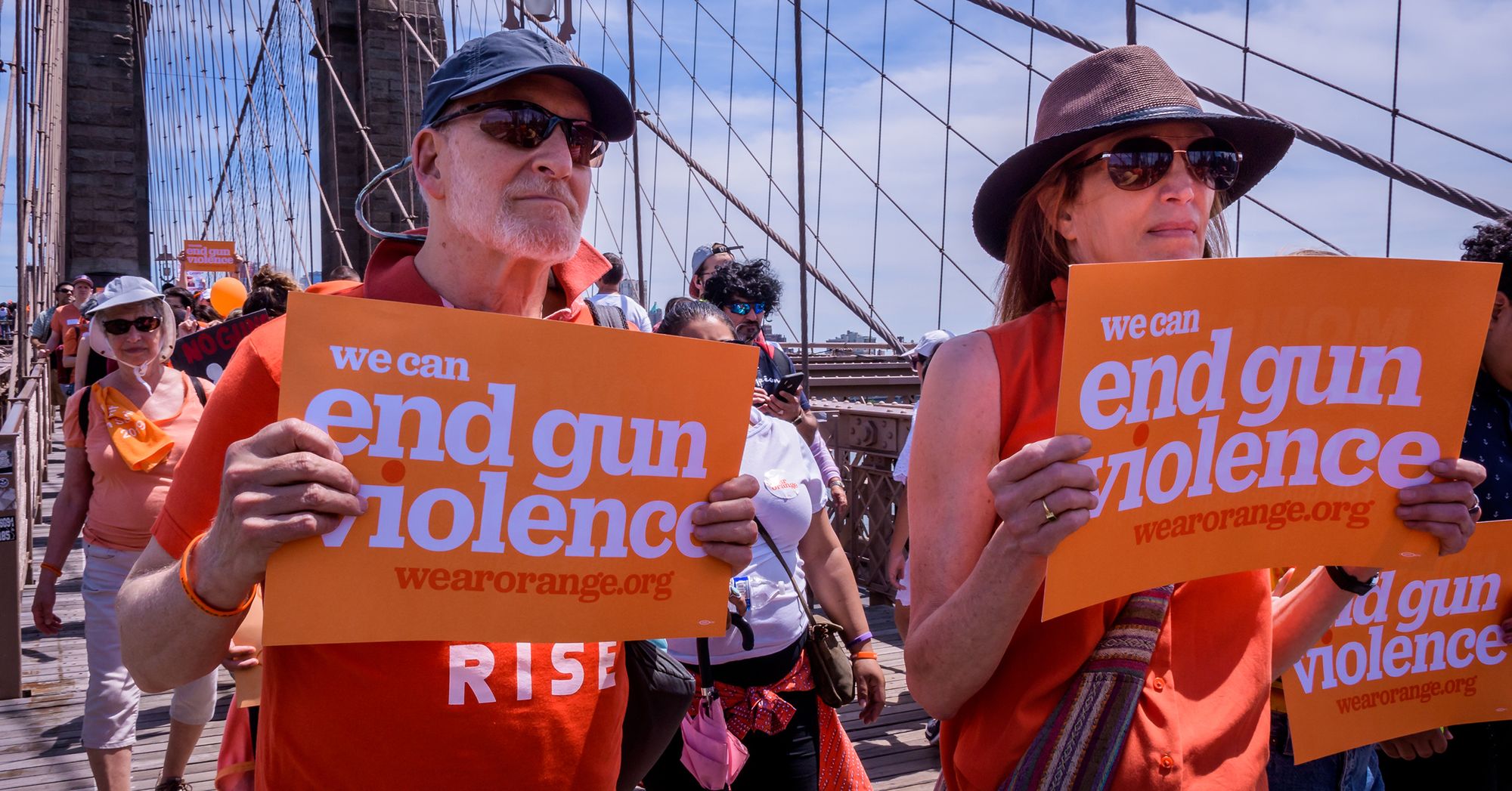 A man with light-toned skin and a woman with light-toned skin carried orange signs reading "We can end gun violence" as people behind them follow during a protest. 