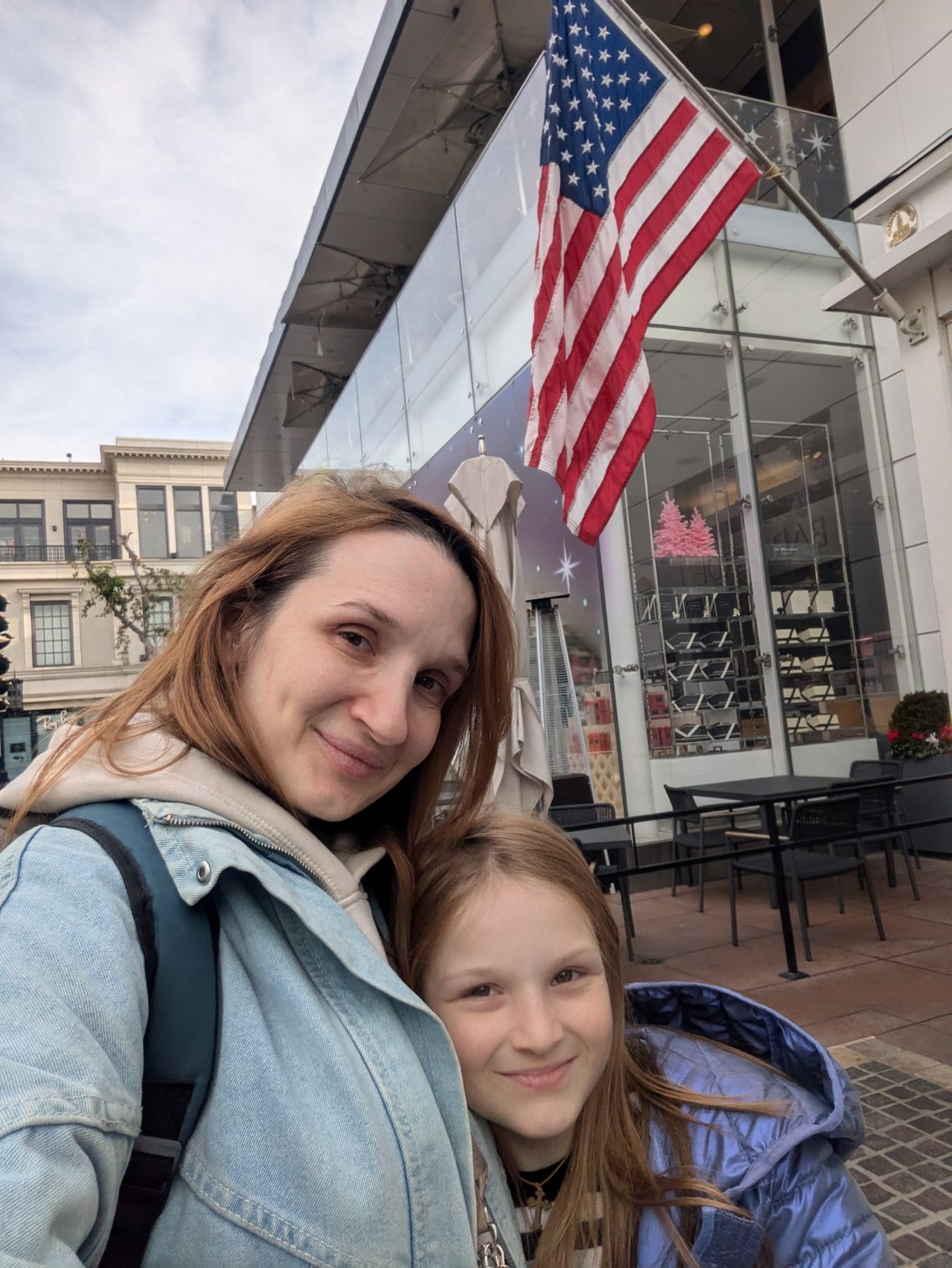 A photo shows a White woman smiling with her young daughter as they stand in front of a building with a U.S. flag. 