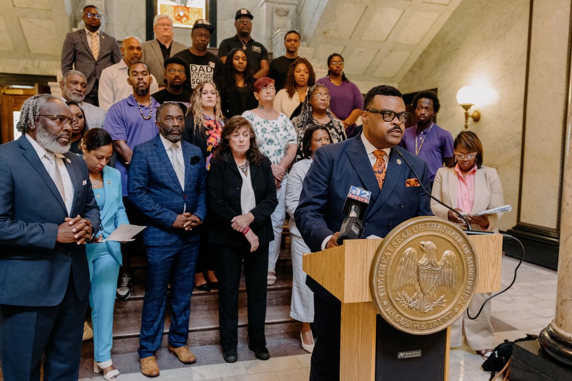 A photo of Kabir Karriem, a Black man, speaking at a podium while wearing a blue suit and glasses. Karriem has short black hair and a mustache. More than 20 people stand behind him. 