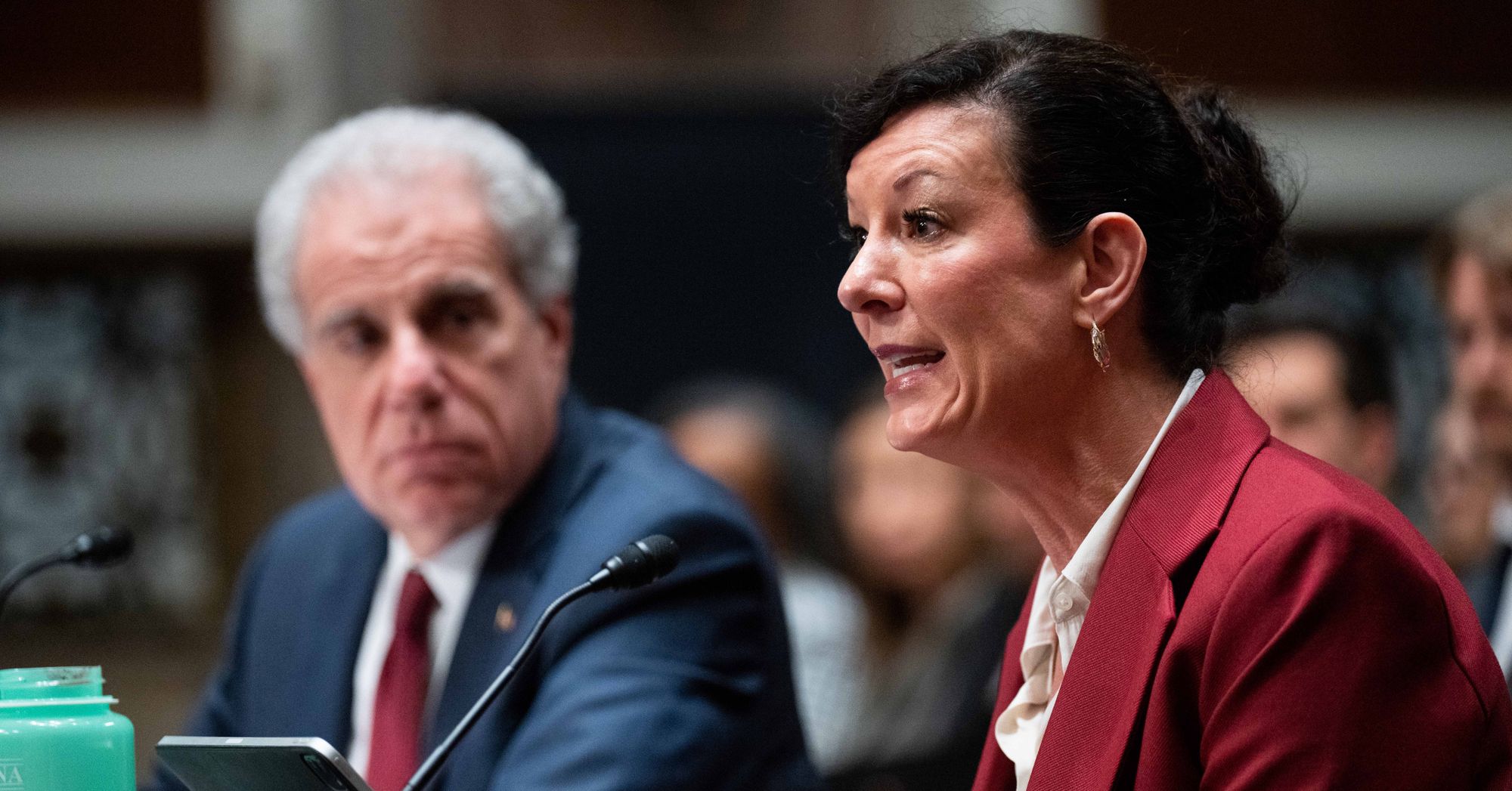 Colette Peters, a White woman in a red suit jacket and short brown hair, speaks, as Michael Horowitz, a White man in a navy suit jacket and white hair, looks on.  