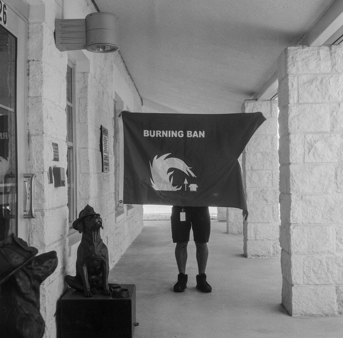 A black-and-white photo of a person holding up a flag with the words “BURNING BAN” above an illustration of a large fire curling over a tree and a building. 