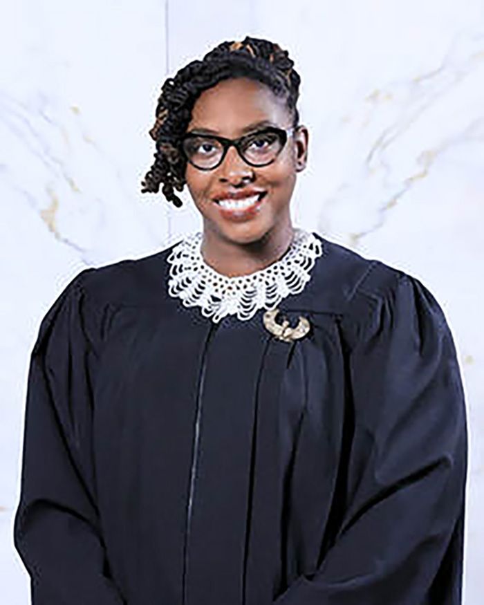 A smiling Black woman is wearing a judge’s robe with a white-beaded collar necklace. 