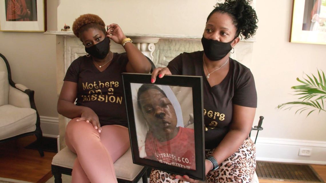 Angela Williams, a Black woman wearing a mask, a black T-shirt and cheetah patterned leggings, sits and holds a framed picture of her son after he was beaten by police officers. A Black woman, wearing a black T-shirt that reads “Mothers on a Mission” and pink leggings, sits next to her.