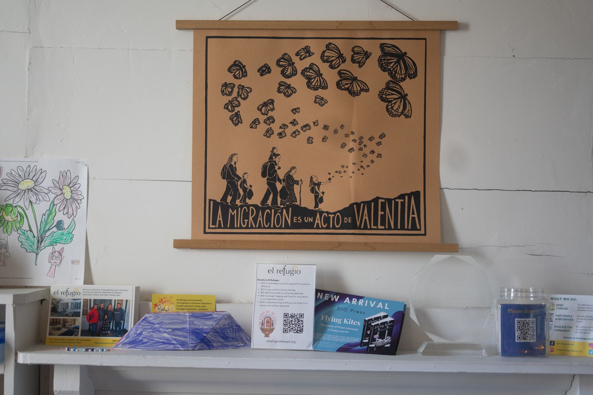 A pale brown sign with black printing that says "La Migración es un acto de Valentia" hangs on a wall. The sign illustrates a group of people with backpacks and walking sticks and a young girl reaching toward a large number of butterflies.