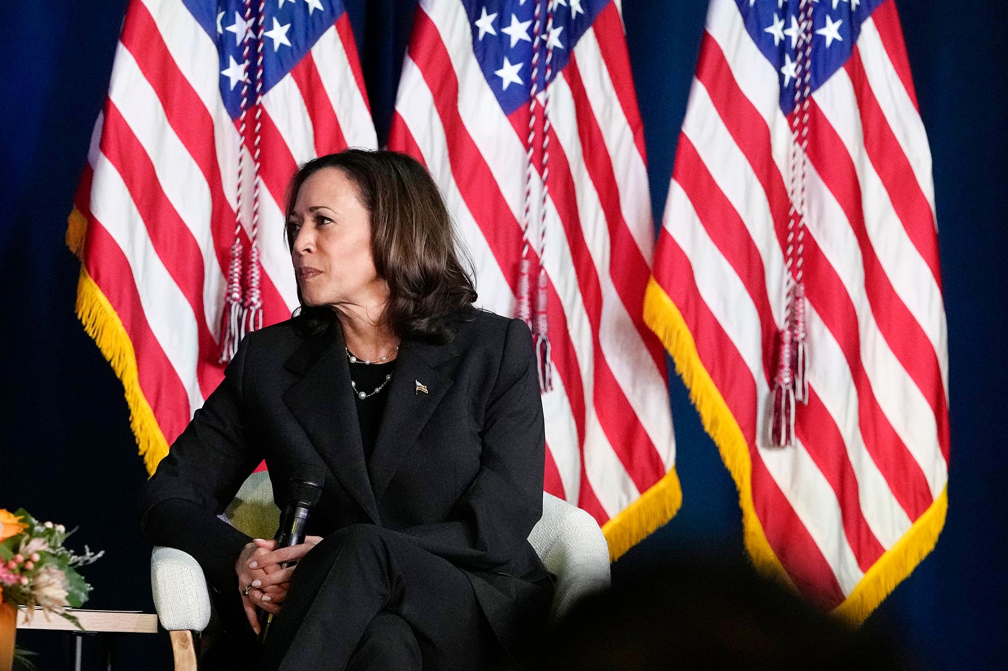 Vice President Kamala Harris, an Indian and Black woman with medium skin tone wearing a black suit, looks to the left while holding a microphone. She is sitting on a chair in front of three American flags. 