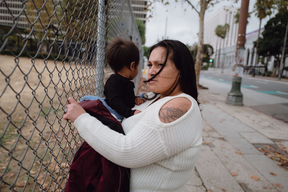 A photo shows Jazmine Mapes, a Native American woman with long dark hair, wearing a white sweater, holding onto a chain link fence with one hand and holding her infant son in the other. 