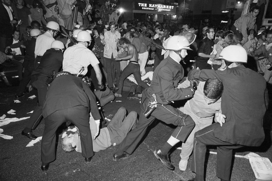 A black-and-white photo shows two police officers struggling to restrain a protester, as police and a group of protesters clash in the background.