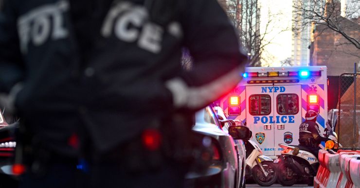 A New York Police Department ambulance is parked on the right, with an officer on a motorcycle behind it. A officer's chest is out of focus in the foreground on the left. 