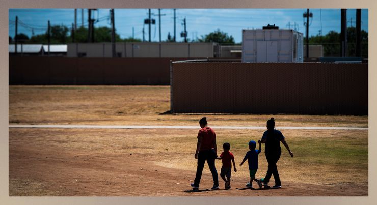 Two women and two children walk together outdoors with their backs facing the camera. Fencing is visible in front of them, along with some rectangular structures and power lines. The grass on the ground is dry.    