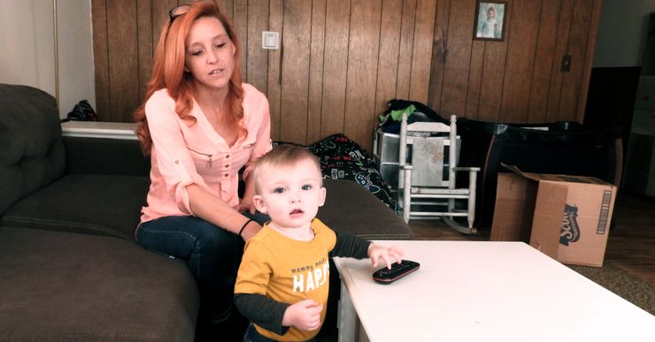 A woman with light-toned skin and reddish long hair sits on a sofa, with a toddler in front of her.  