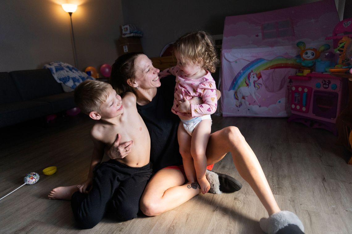 A photo of a White woman sitting on the floor while smiling and hugging her two children.