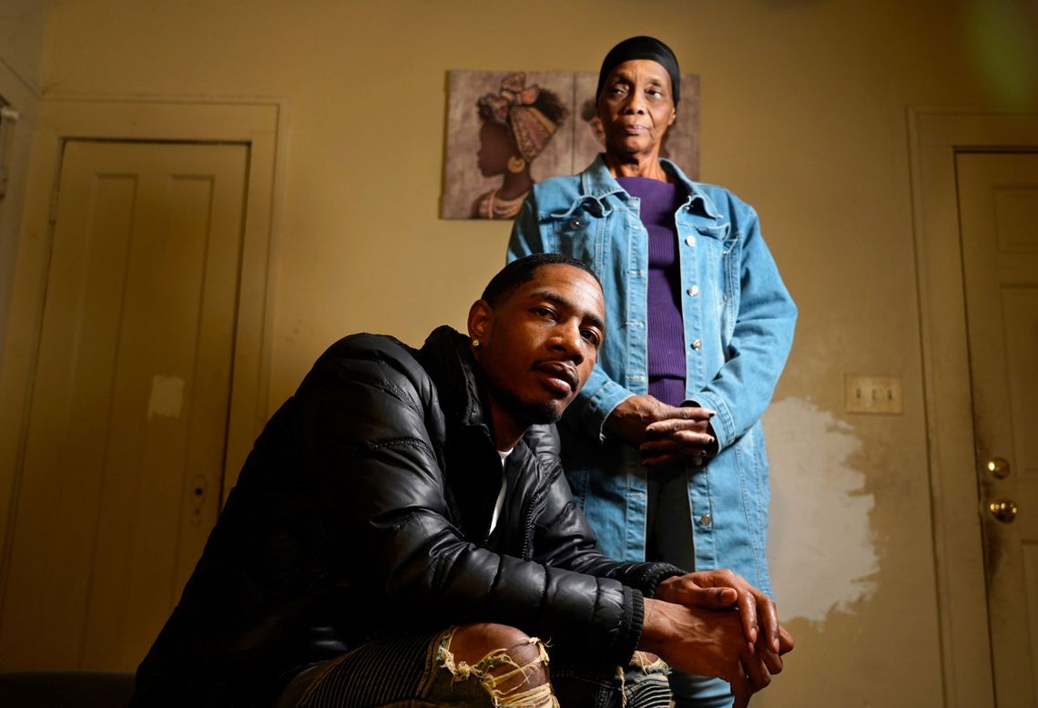 A Black man and his grandmother pose for a portrait in their apartment. 