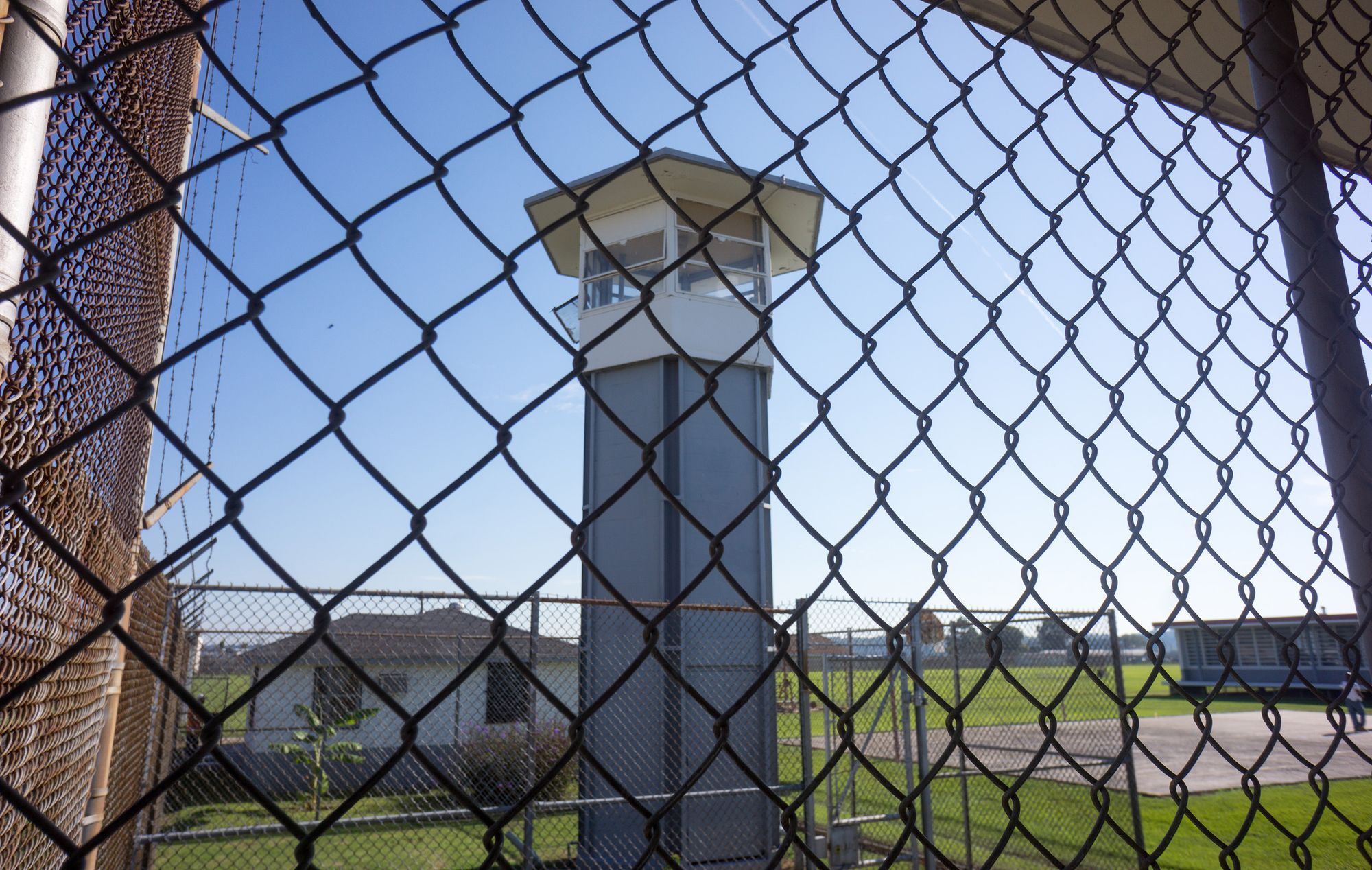 A prison tower and grounds are seen behind a chain-link fence in the foreground.  