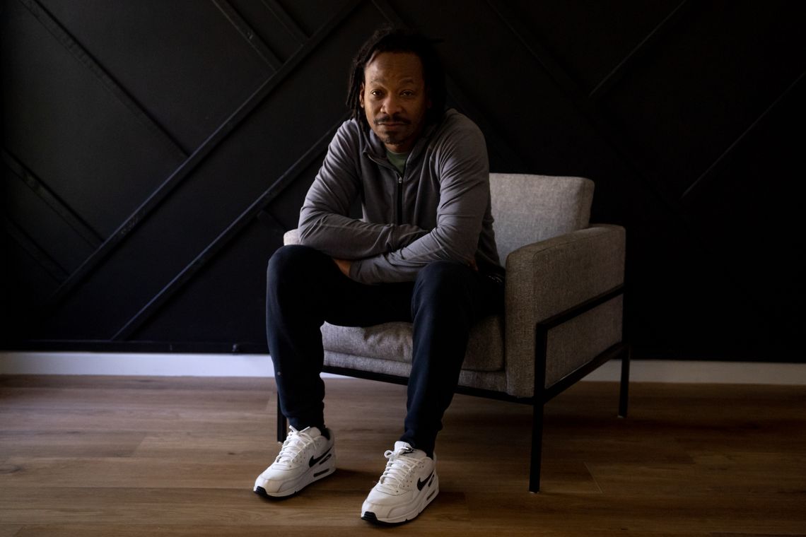 A Black man wearing a gray pullover, navy sweatpants and white sneakers sits on a chair at his home.