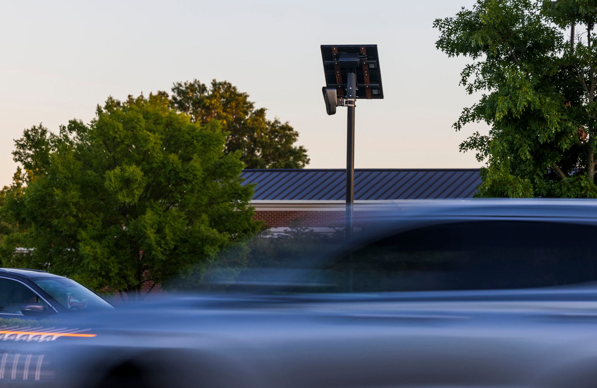 A photo shows cars driving in a blur past a Flock camera, which is situated at the top of a pole. There is a square plate at the top of the pole. Behind the camera are some trees and a building. 