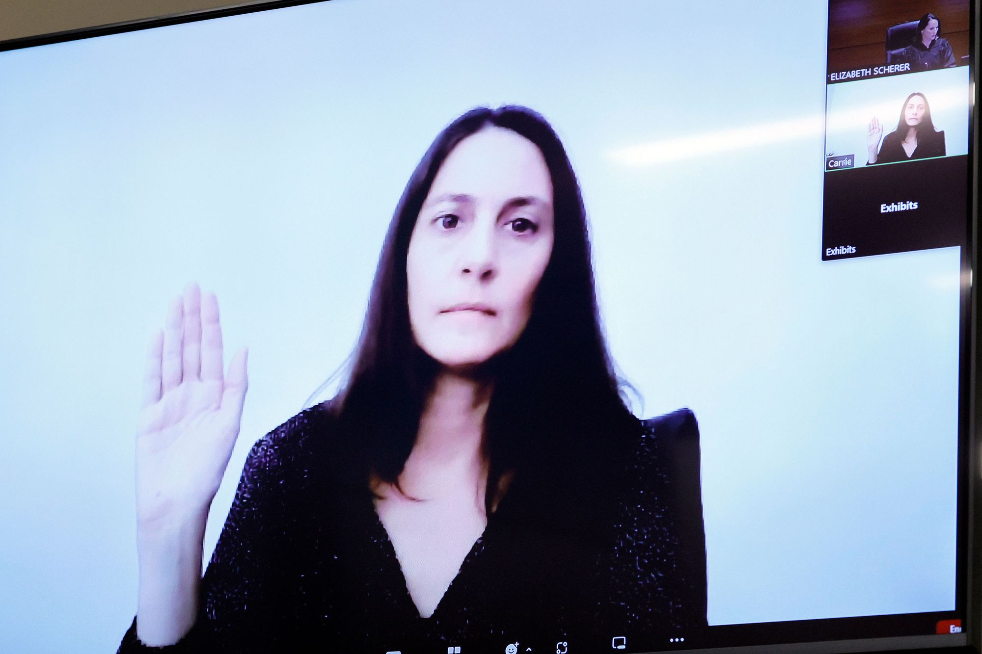 A photo of a White woman with dark hair raising her right hand, seen through a video call projection. 