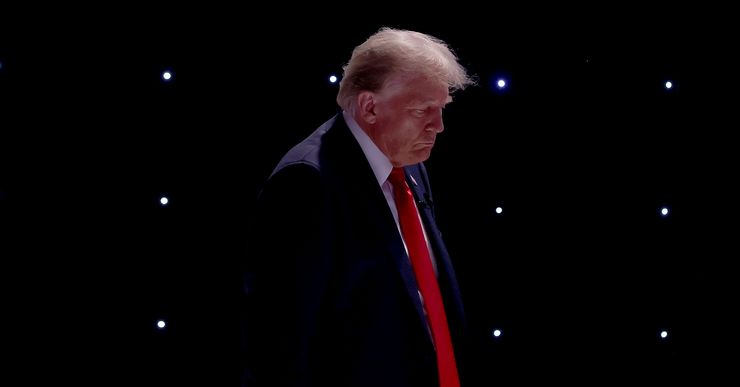 Former President Donald Trump, a White man wearing a black suit and red tie, looks down. There are small studio lights in the background. 