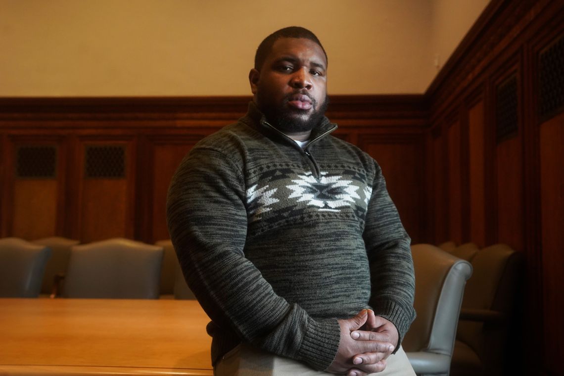 A Black man in a dark green patterned sweater sits on the edge of a conference room table and looks into the camera. 