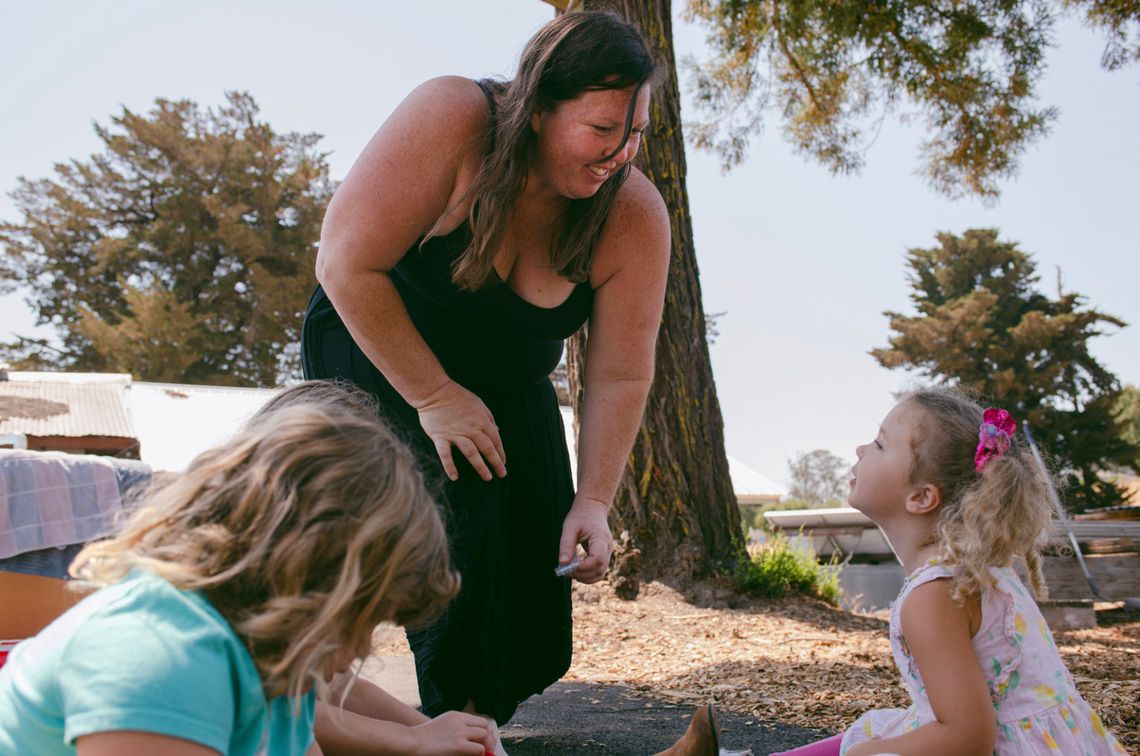 Susan Horton, a White woman wearing a black tank top and black pants, leans while she stands to talk to one of her daughters. In the foreground, another daughter, wearing a light blue shirt, is looking at the floor. 