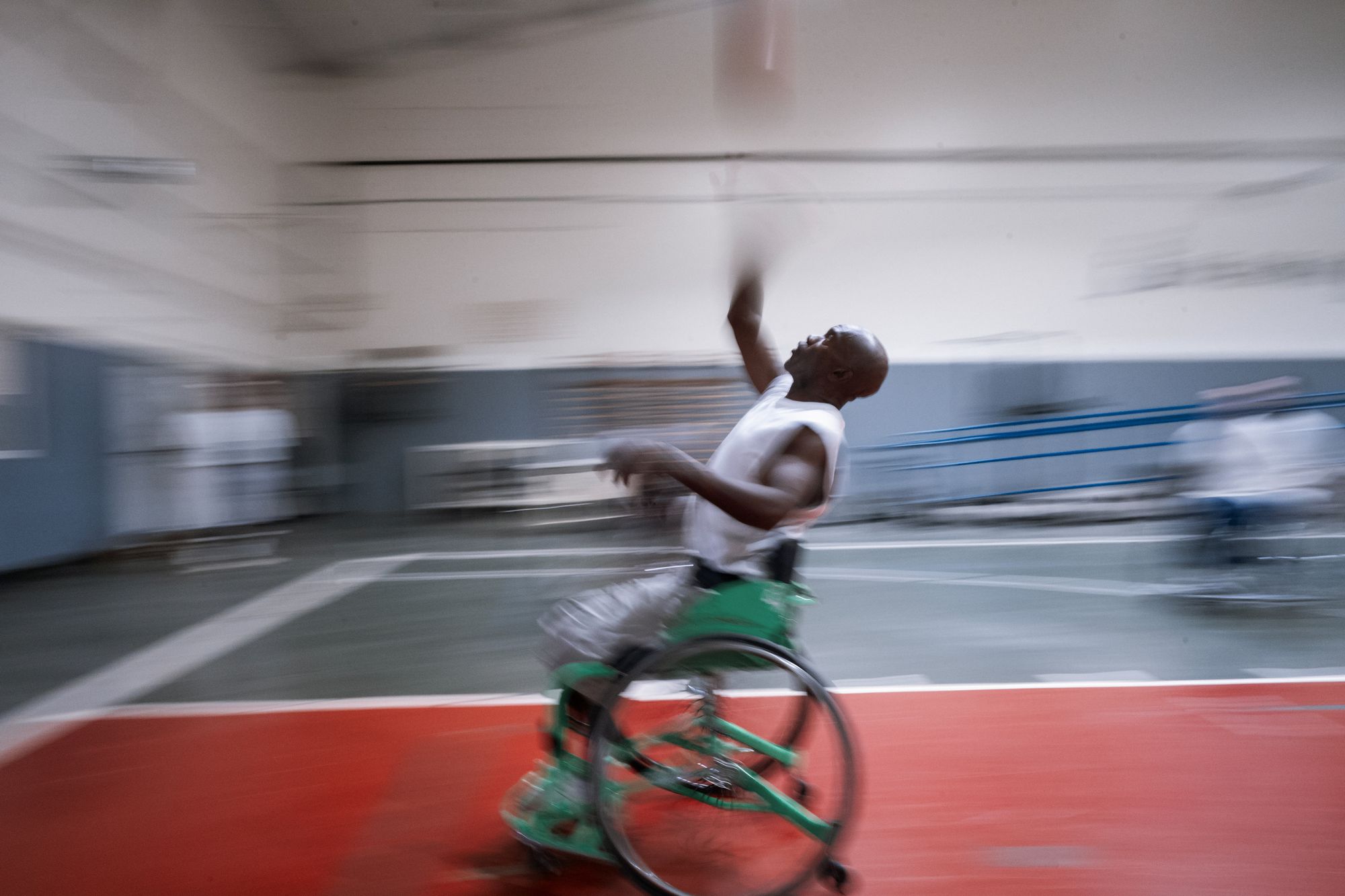 A photo shows a Black man speeding across the court in a blur in a wheelchair while reaching up for a basketball. 