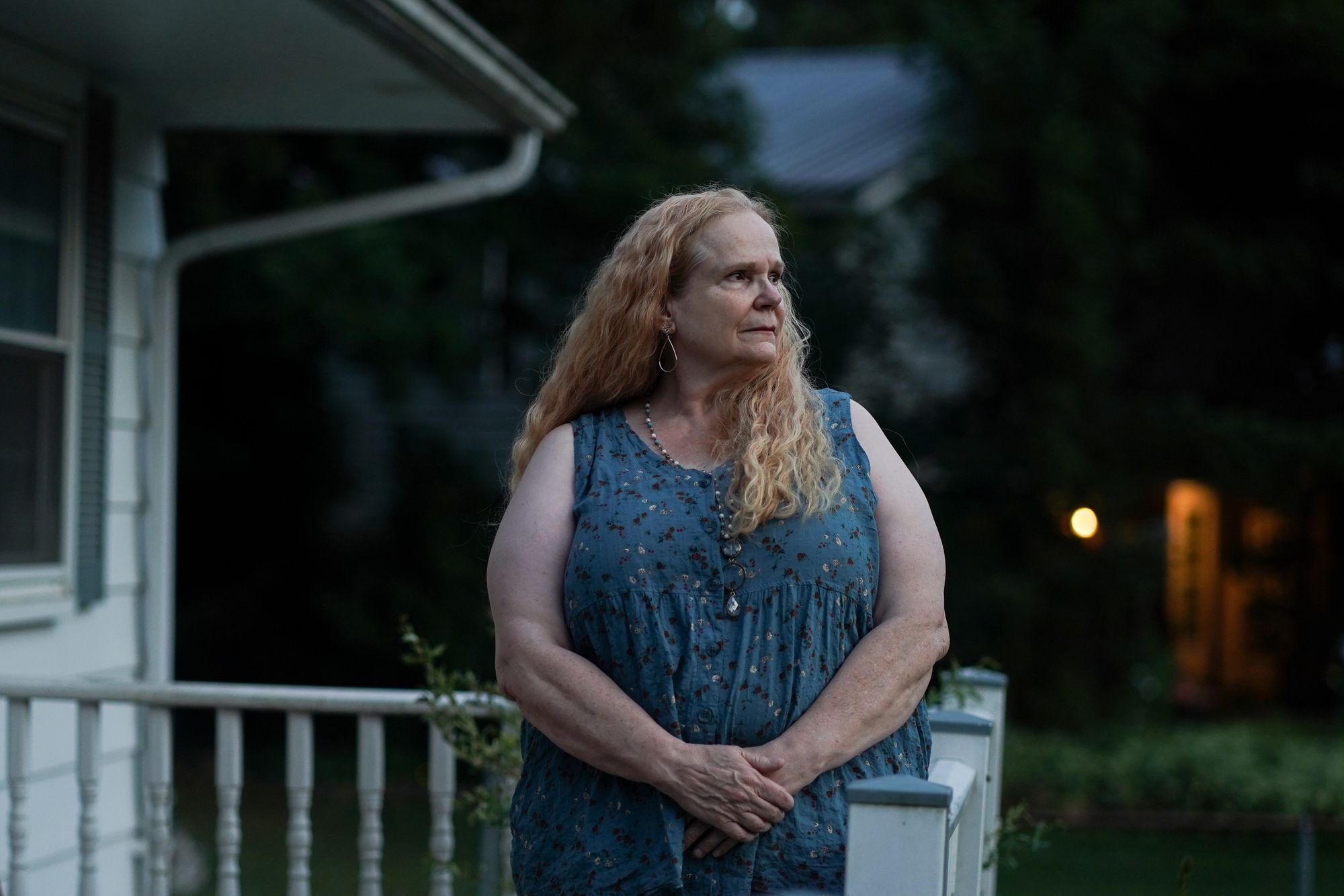 A White woman with wavy blonde hair stands on her porch at dusk.  