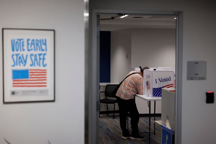 A photo shows a person, wearing a hat, a red, green, and blue flannel shirt, and black pants, voting in a voting booth with the American flag printed on it.