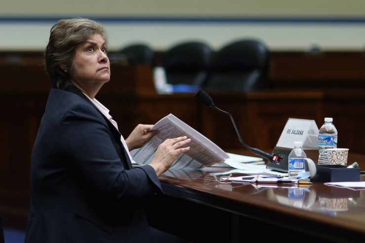 A photo of a Latina woman in her 60s with a medium skin tone and short hair sitting at a desk with a microphone. The woman is wearing a blazer and blouse while holding a stack of papers, and is looking up and to the side. 