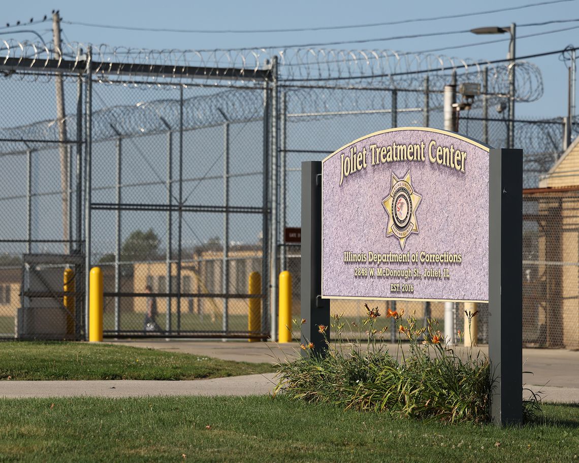A photo shows a sign on a lawn that reads, “Joliet Treatment Center.” There are yellow buildings surrounded by barbed wire fencing in the background. 