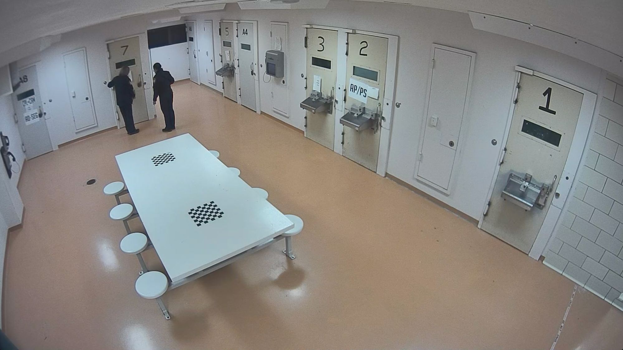 Two still images from a surveillance camera show a pod of jail cells. In the first image, the pod is empty. In the second image, two correctional officers are standing outside a door with the number “7.” 