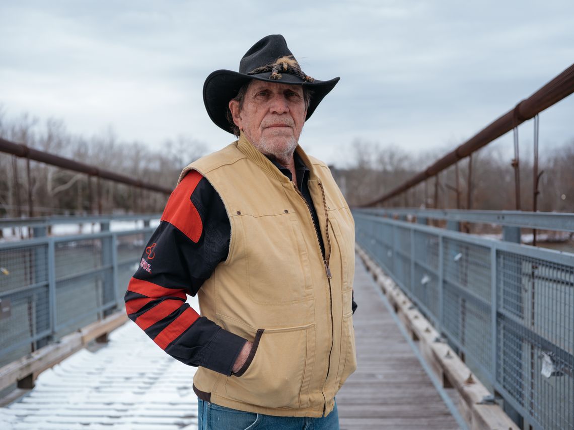 George Hall, an older White man wearing a hat with a brim and a tan vest and long sleeved jacket, stands on a bridge.