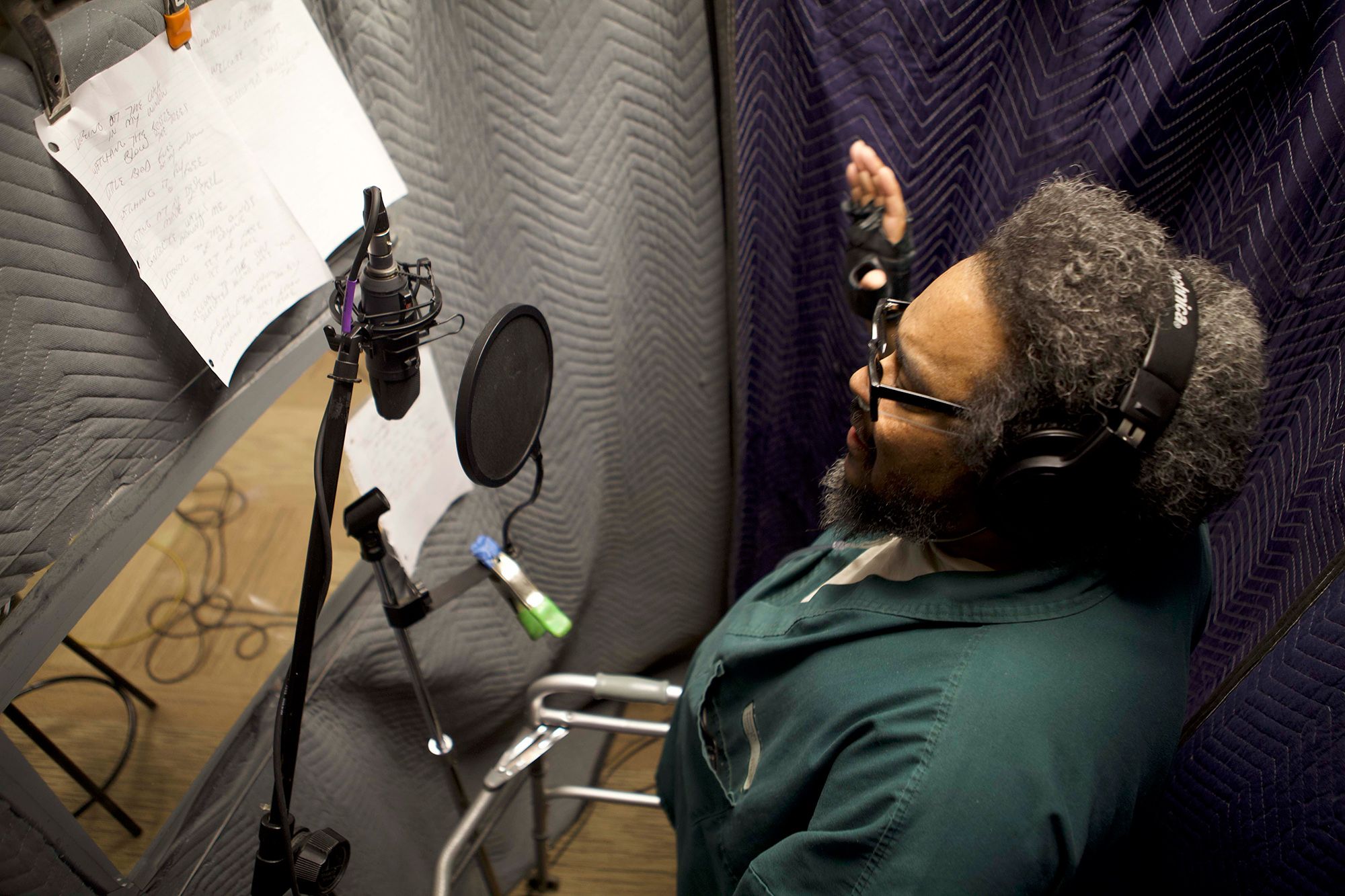 A man inside the recording studio looks at a page of song lyrics taped to the interior wall as he sings into a microphone.